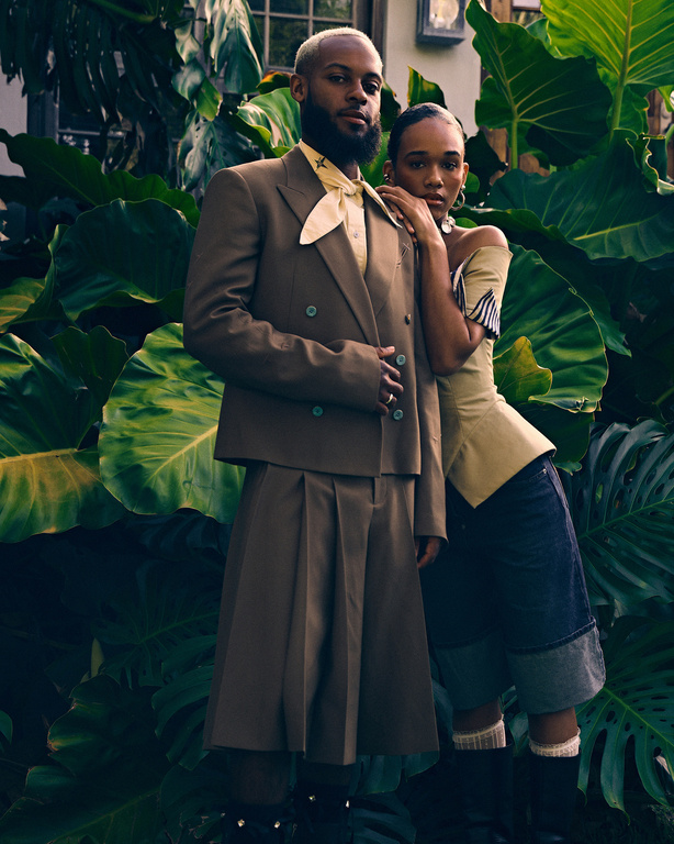 A stylish couple poses outdoors amid lush greenery, the man in a brown suit and kilt, the woman in a yellow top and denim shorts.