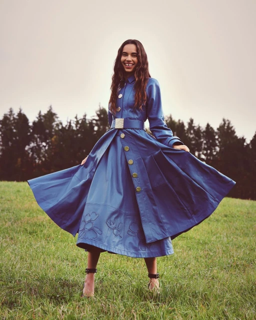 A happy woman in a blue belted dress and high heels poses with arms outstretched in a green meadow with a tree line in the background.