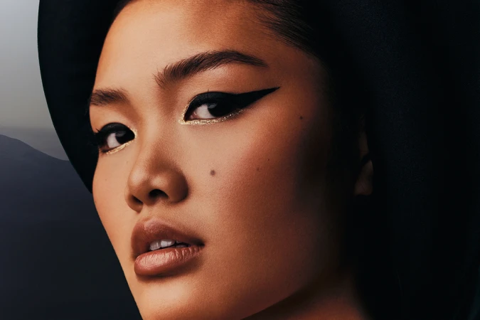 Close-up of an Asian woman with dramatic eye makeup and a hat, against a blurred background featuring a mountain range.