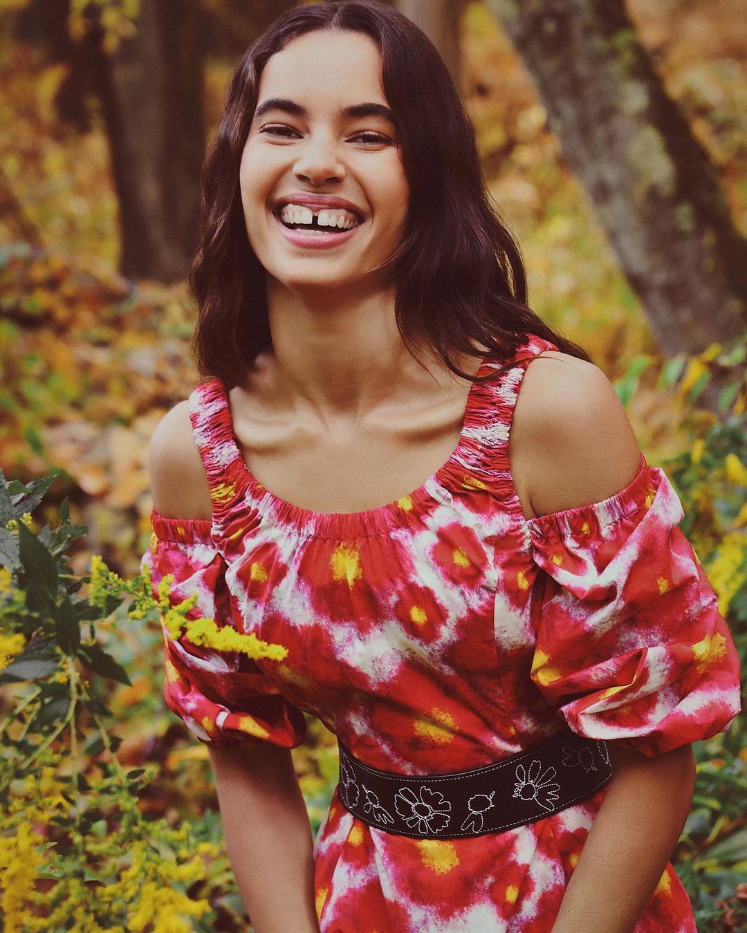 Portrait of a smiling brunette woman with a gap-toothed smile wearing a red and white patterned off-the-shoulder dress in a forest setting.