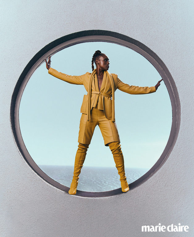 A woman poses in yellow-gold suit and matching thigh-high boots inside a large circular opening, with the ocean and sky visible behind.