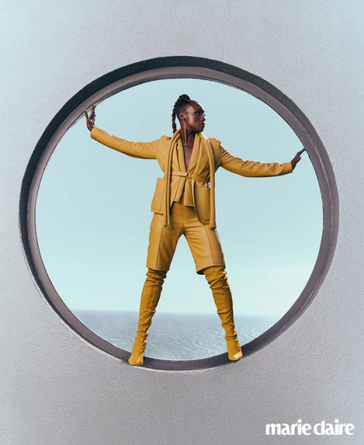 A woman poses in yellow-gold suit and matching thigh-high boots inside a large circular opening, with the ocean and sky visible behind.