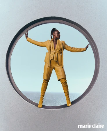 A woman poses in yellow-gold suit and matching thigh-high boots inside a large circular opening, with the ocean and sky visible behind.