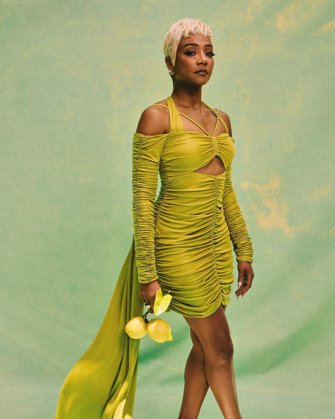 Tiffany Haddish poses in a green dress, holding lemons against a green backdrop, showcasing a blend of fashion and nature-inspired elements.