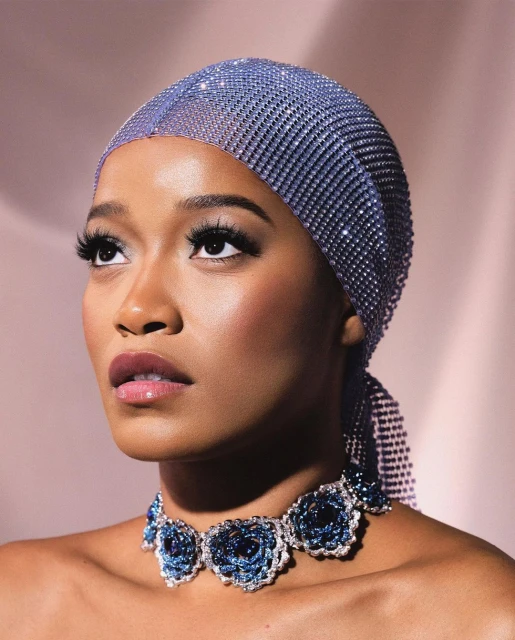 Close-up of a stylish African-American woman wearing a sparkly mesh headscarf and blue floral necklace, looking upwards against a pink background.