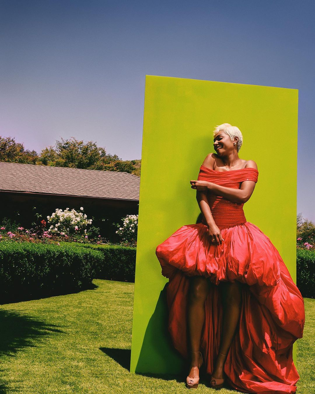 Woman in a red off-the-shoulder gown poses next to a vibrant green wall in a sunlit outdoor setting with a clear blue sky and lush lawn.
