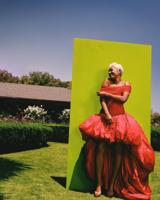 Woman in a red off-the-shoulder gown poses next to a vibrant green wall in a sunlit outdoor setting with a clear blue sky and lush lawn.