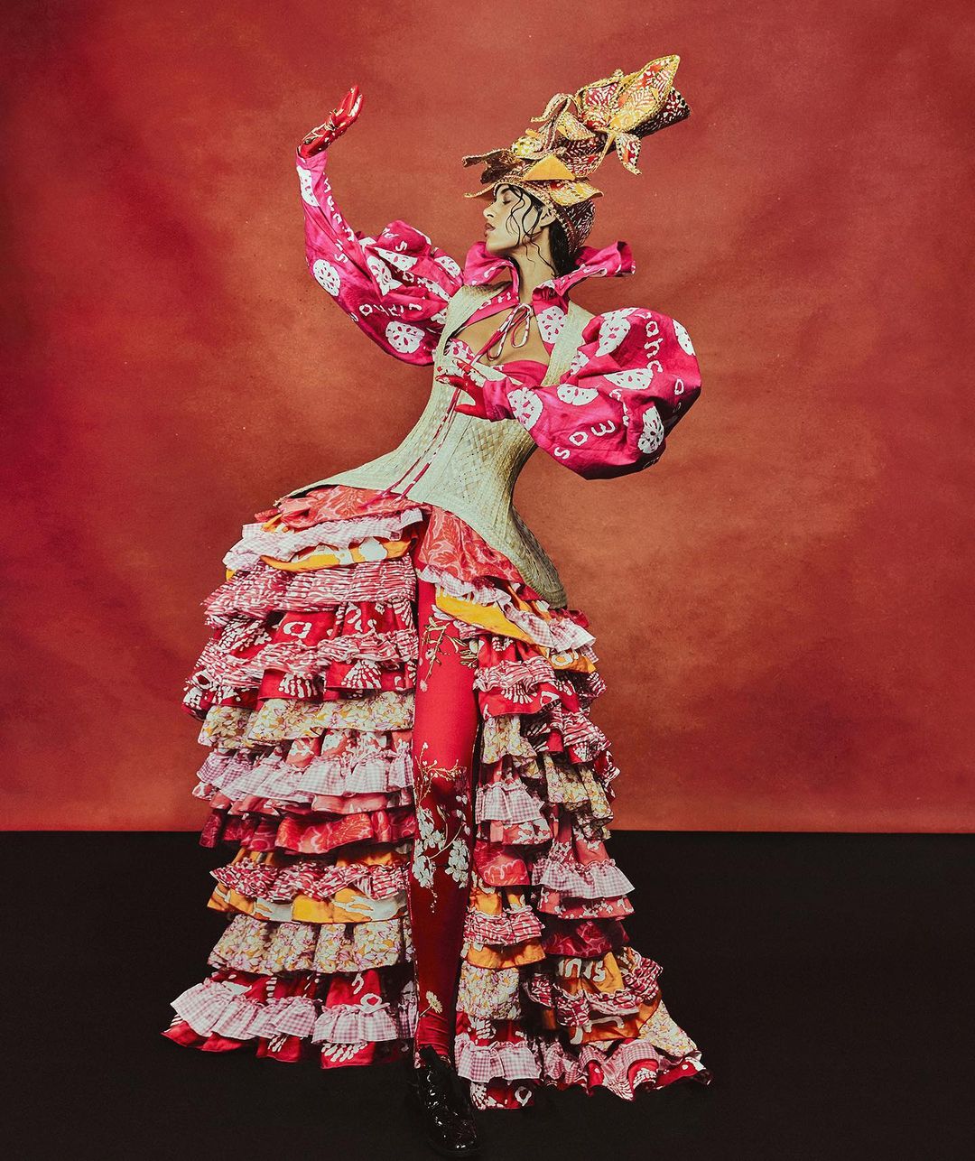 Fashion model poses in a red and white dress with layered ruffles, complemented by an elaborate headpiece on a red background.