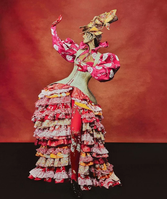 Fashion model poses in a red and white dress with layered ruffles, complemented by an elaborate headpiece on a red background.