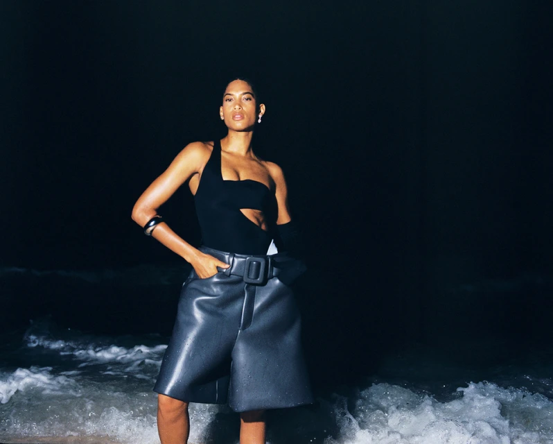 Fashion shot of a woman in a one-shoulder top and leather shorts standing on a beach at night with visible ocean waves.
