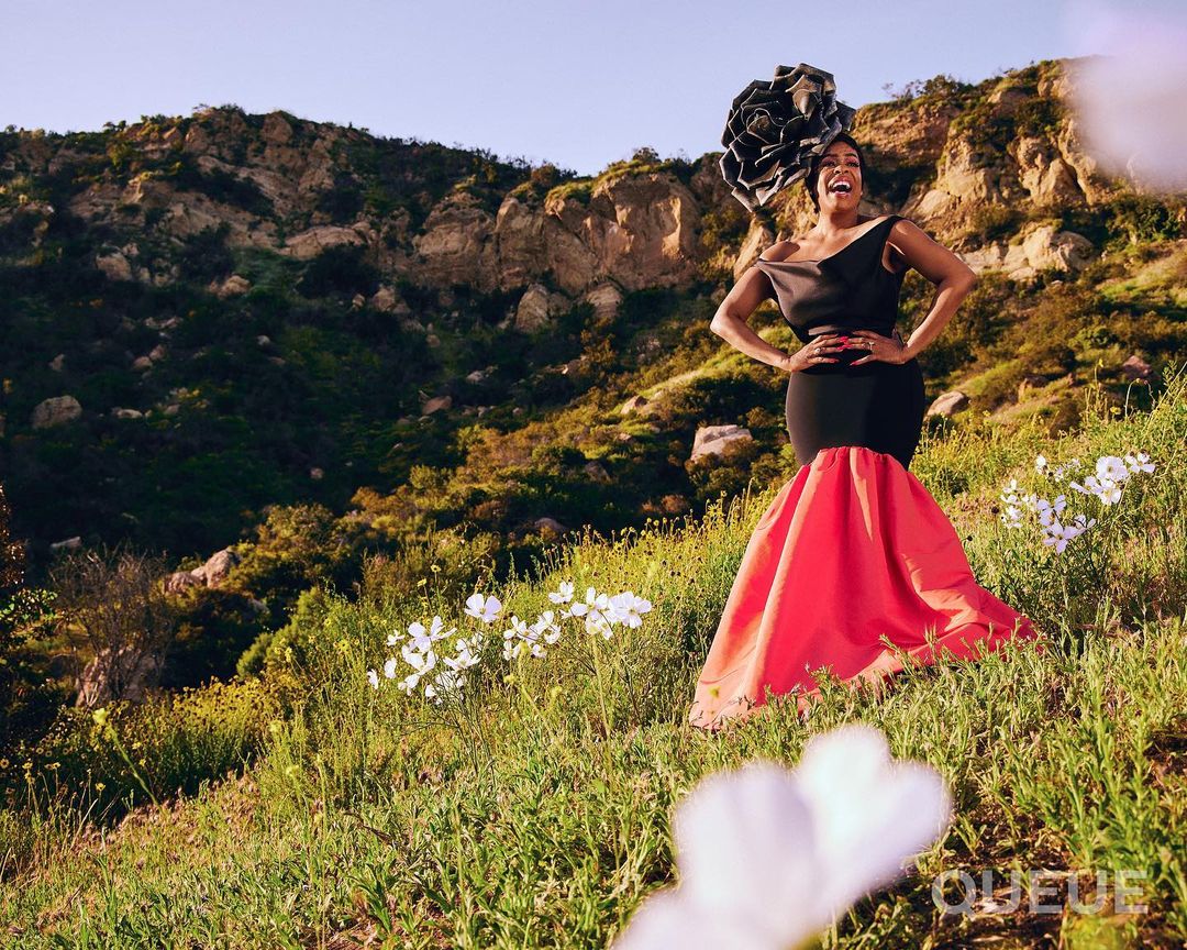 Elegant woman in a black and coral gown poses in a grassy field with white flowers and a mountain backdrop, showcasing fashion.