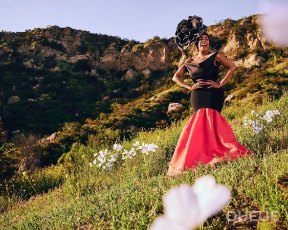 Elegant woman in a black and coral gown poses in a grassy field with white flowers and a mountain backdrop, showcasing fashion.