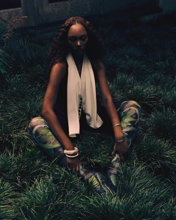 Woman with curly brown hair seated in tall grass wearing patterned leggings and a white top