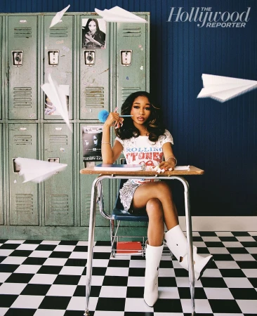 Normani poses at a school desk in front of green lockers with paper airplanes, wearing a Rolling Stones t-shirt and white boots.