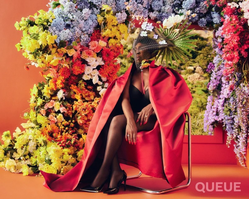 Woman in black top and red cloak sitting on chair, surrounded by colorful flowers against an orange backdrop, looking elegant.