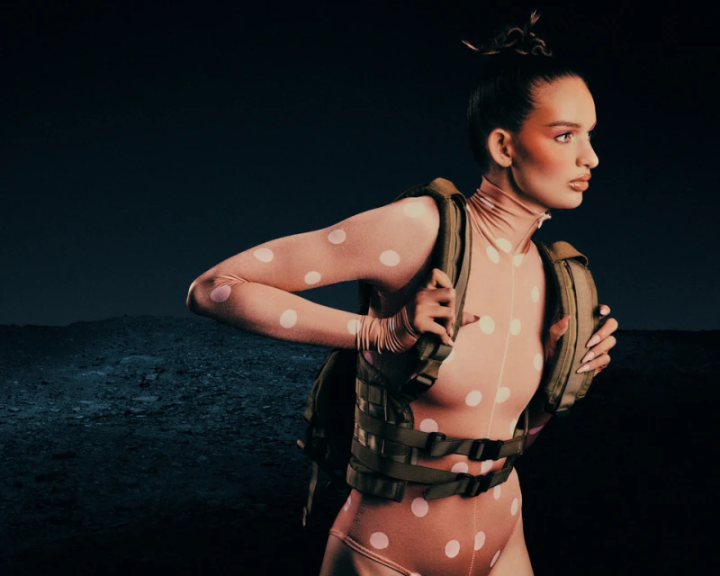 Woman in pink polka dot bodysuit and tactical vest poses outdoors against a dark backdrop with textured ground under studio lighting.