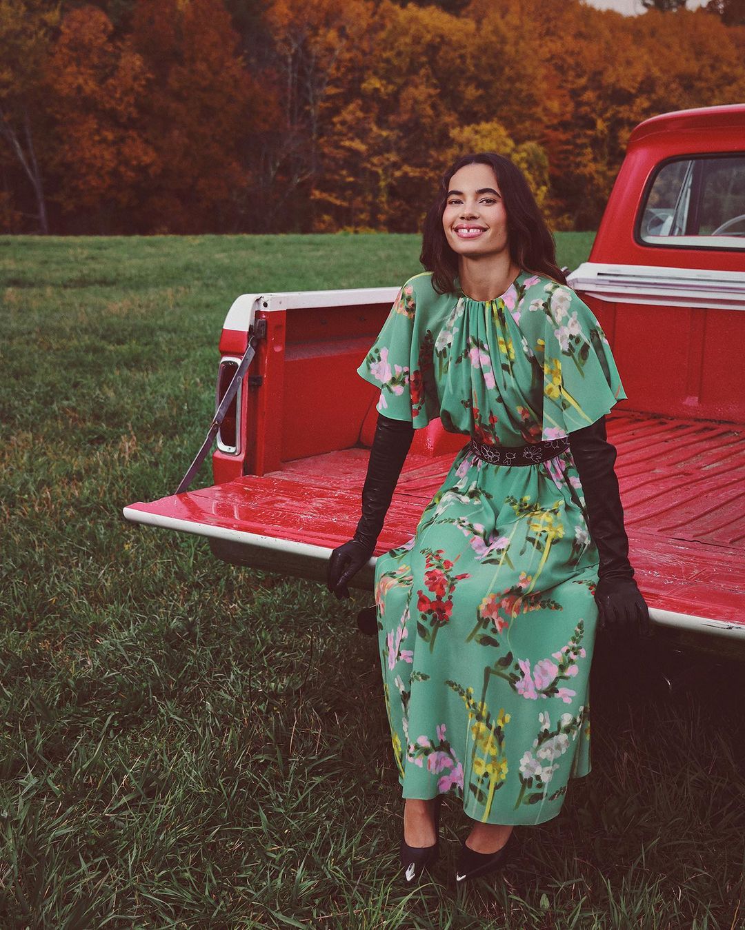 A woman wearing a green floral dress sitting on the open tailgate of a red pickup truck in a grassy field with an autumn forest backdrop.