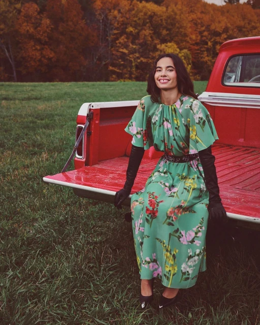 A woman wearing a green floral dress sitting on the open tailgate of a red pickup truck in a grassy field with an autumn forest backdrop.