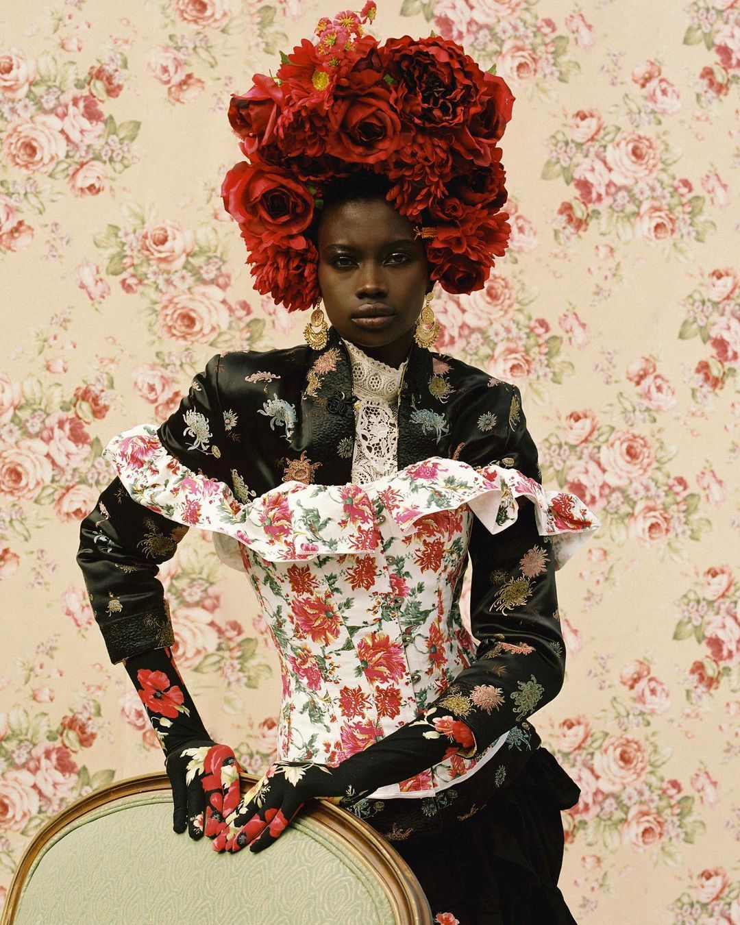 Elegant portrait of a Black woman in floral attire and red rose headpiece against a floral wallpaper backdrop in a studio setting.