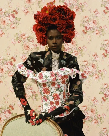 Elegant portrait of a Black woman in floral attire and red rose headpiece against a floral wallpaper backdrop in a studio setting.