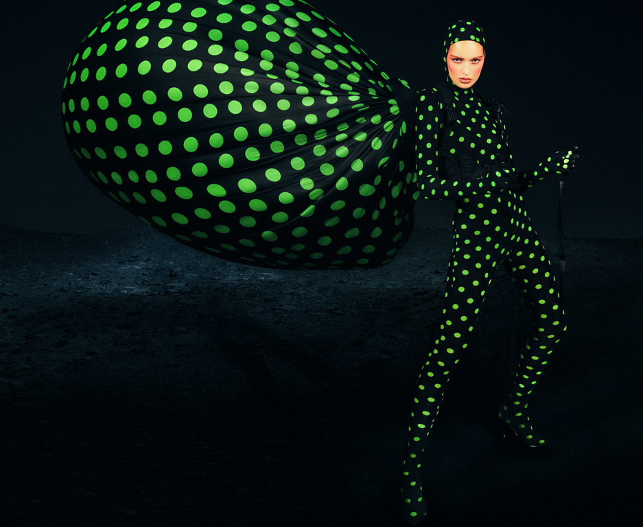 Fashionable woman in full polka dot bodysuit and headscarf poses in a dimly lit space next to an oversized matching sack.