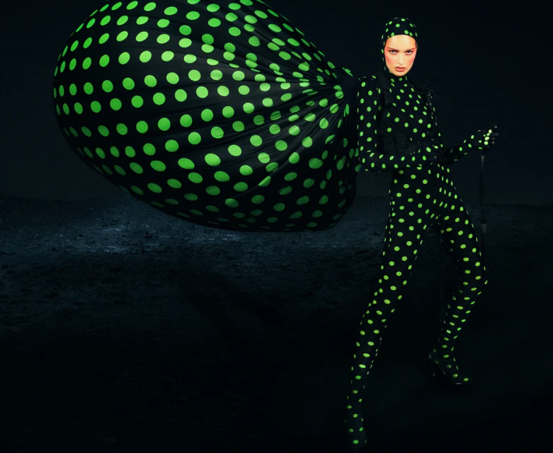 Fashionable woman in full polka dot bodysuit and headscarf poses in a dimly lit space next to an oversized matching sack.