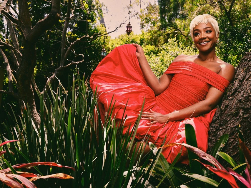 Tiffany Haddish poses in a vibrant red off-the-shoulder dress in a lush outdoor setting with green foliage and dappled sunlight.