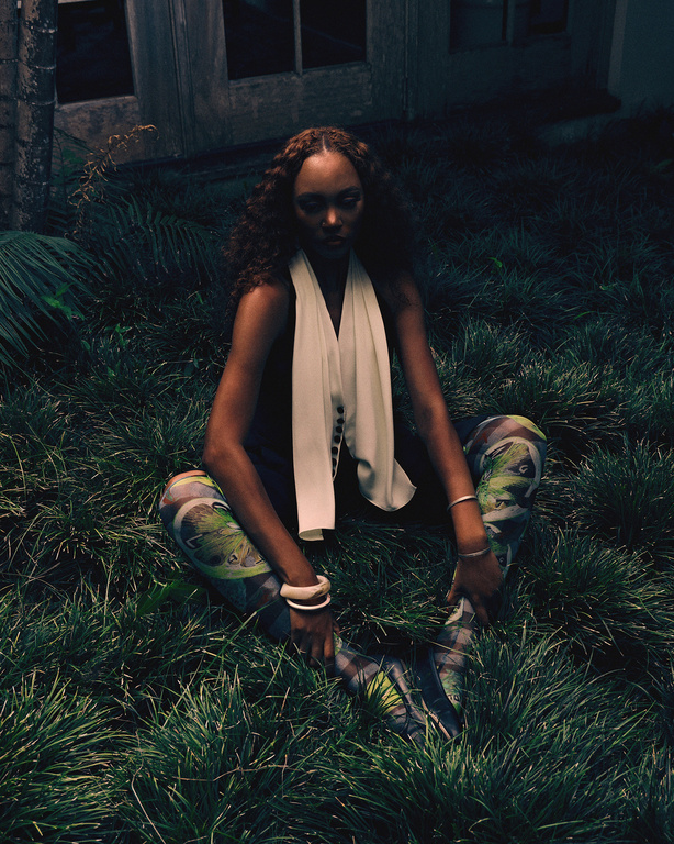 Black woman with curly hair wearing patterned leggings and a white scarf seated outdoors in lush green grass.