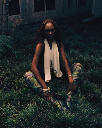 Black woman with curly hair wearing patterned leggings and a white scarf seated outdoors in lush green grass.