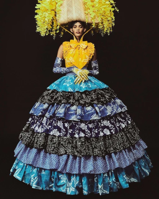 A woman poses in a tiered floral gown and a hat made of flowers against a black background, showcasing a unique fashion statement.