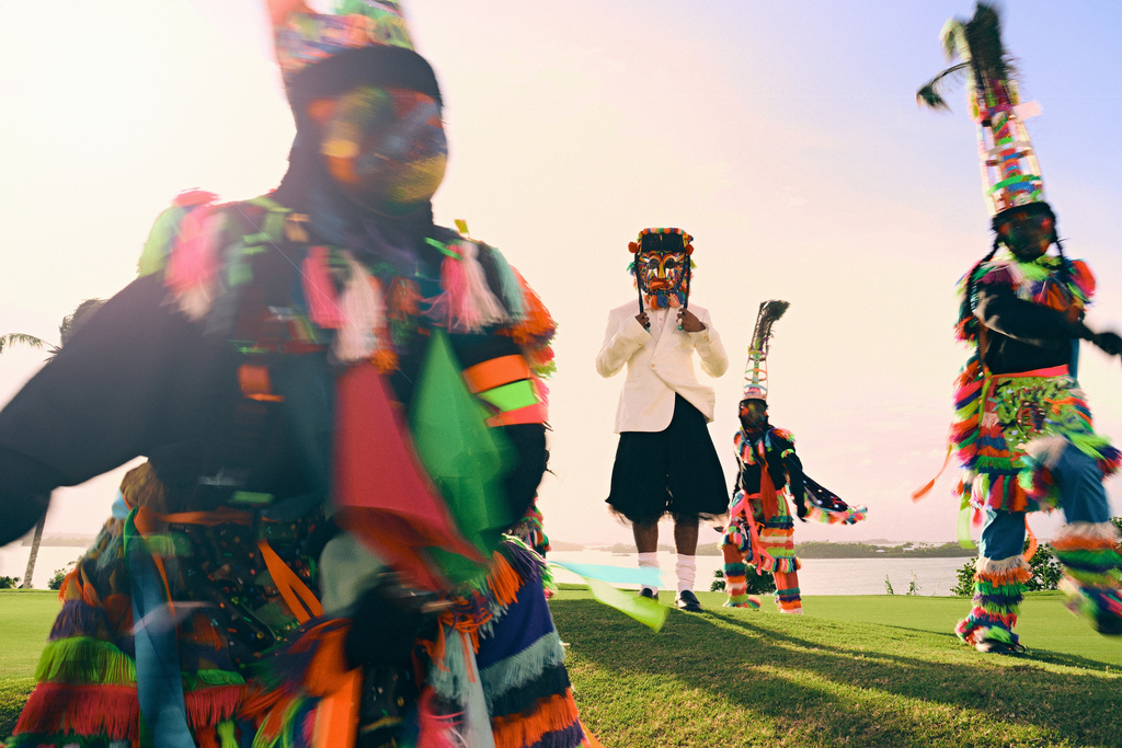 A group of Junkanoo performers in vibrant costumes, including one in a white suit, parade on a grassy field with a coastal view in the background.