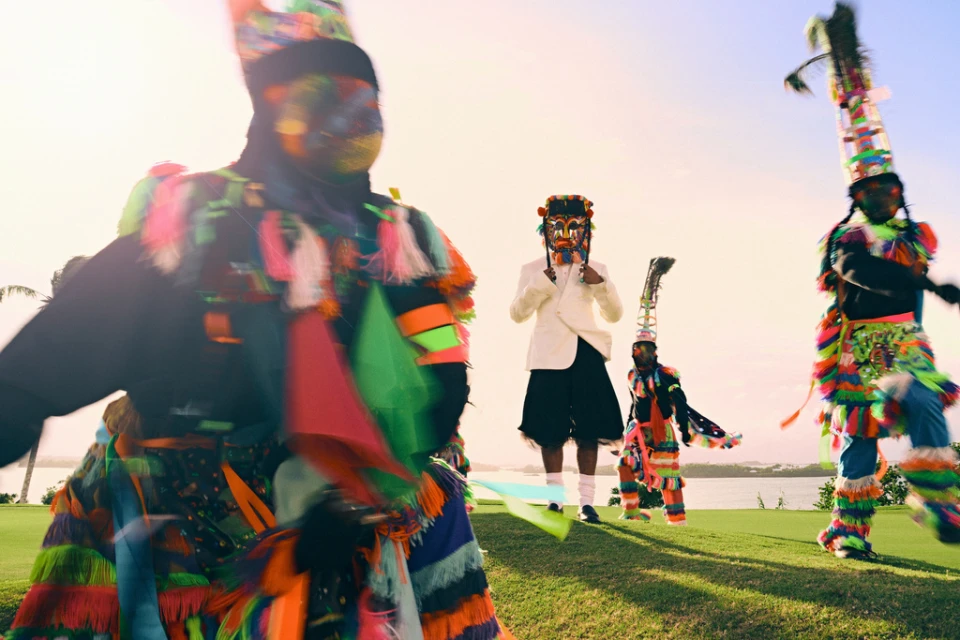 A group of Junkanoo performers in vibrant costumes, including one in a white suit, parade on a grassy field with a coastal view in the background.