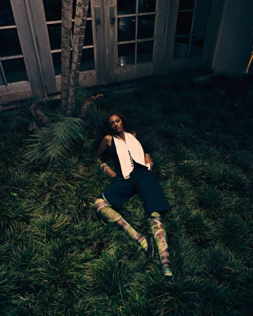 A woman lying on a lush green lawn wearing dark trousers, a white vest, and patterned leggings, photographed from above.