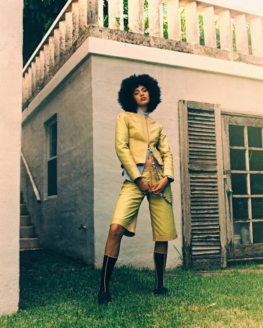 Woman with afro poses in yellow jacket and shorts outside white building. Dark socks, grassy lawn, warm light, fashion portrait.
