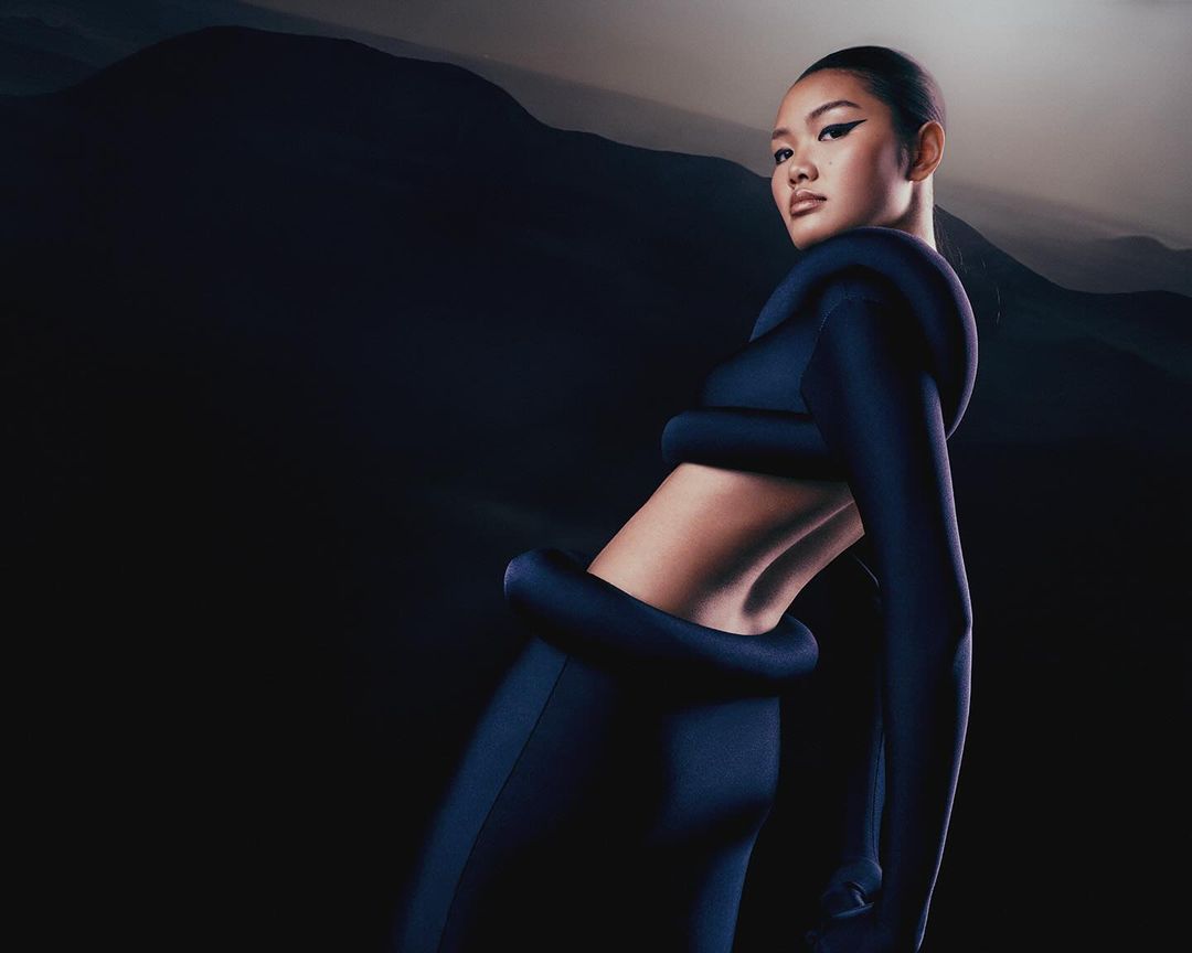 Glamour shot of Asian female model wearing stylish blue outfit against a mountain backdrop, showcasing fashion and scenic beauty.