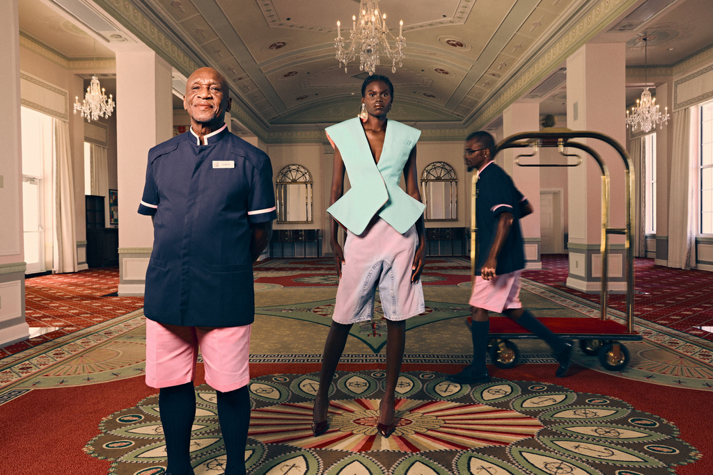 Fashion editorial shot indoors with three people, including a model, bellhop, and man pulling luggage in a hotel lobby with patterned carpet.