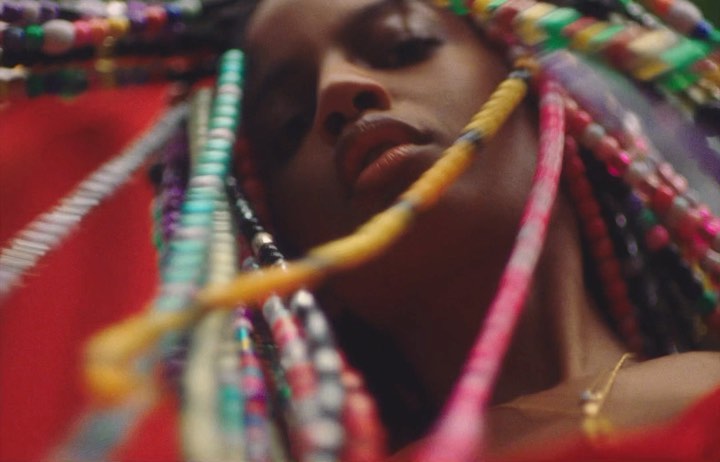 Close-up of a woman with multicolored braids and a gold necklace, with a blurred red and green background.