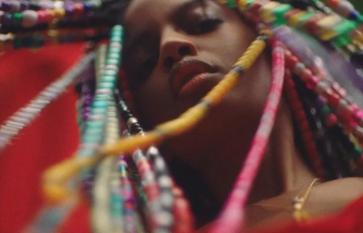 Close-up of a woman with multicolored braids and a gold necklace, with a blurred red and green background.