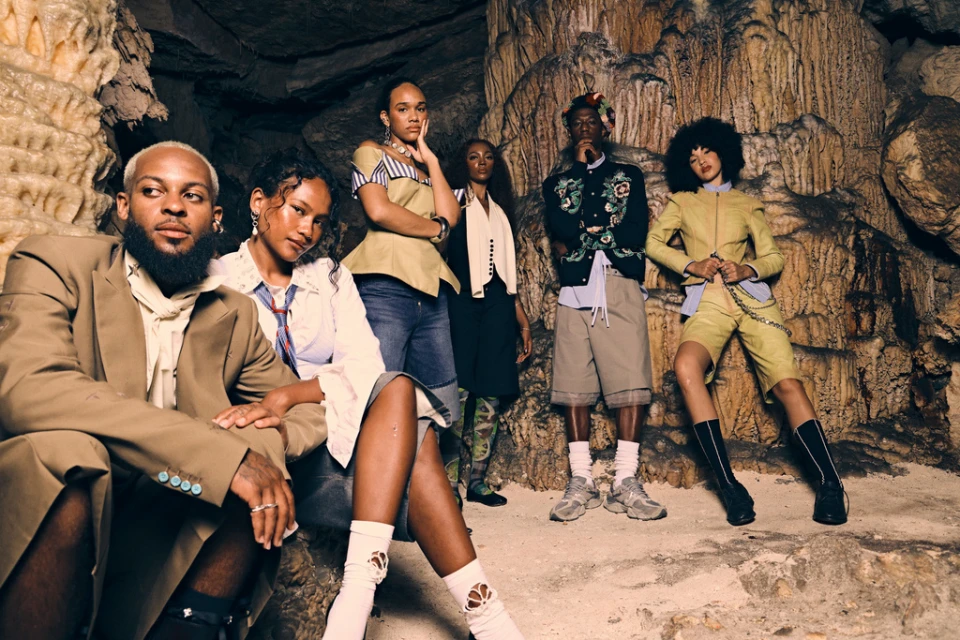 Group of six diverse fashion models in stylish clothing pose inside a cave with textured rock formations and earth-tone colors, shot with medium framing.