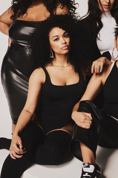 A group of plus-size women pose in a studio wearing black bodysuits, dresses and thigh-high stockings; black, white, and grey are dominant.