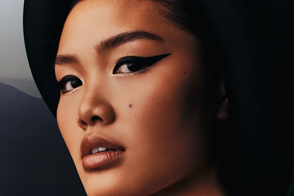 Close-up portrait of a young Asian woman with black eyeliner and hat, showcasing a beauty shot with dramatic lighting.