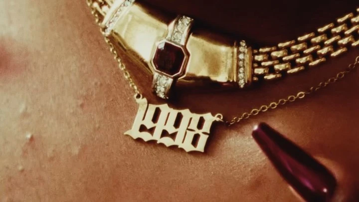 Close-up shot of a gold chain necklace with 1998 pendant, resting on tan skin with a long burgundy fingernail in view.