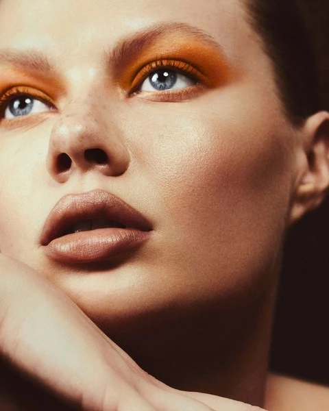 Close-up portrait of a woman with orange eyeshadow and blue eyes, gazing upwards with her face resting on her hand.