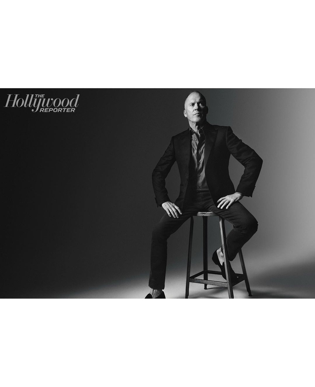 Black and white portrait of a man in a suit sitting on a stool in front of a dark backdrop, with the Hollywood Reporter logo.