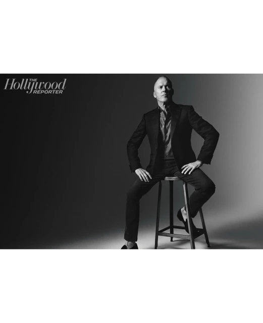Black and white portrait of a man in a suit sitting on a stool in front of a dark backdrop, with the Hollywood Reporter logo.