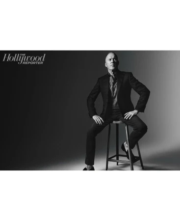 Black and white portrait of a man in a suit sitting on a stool in front of a dark backdrop, with the Hollywood Reporter logo.