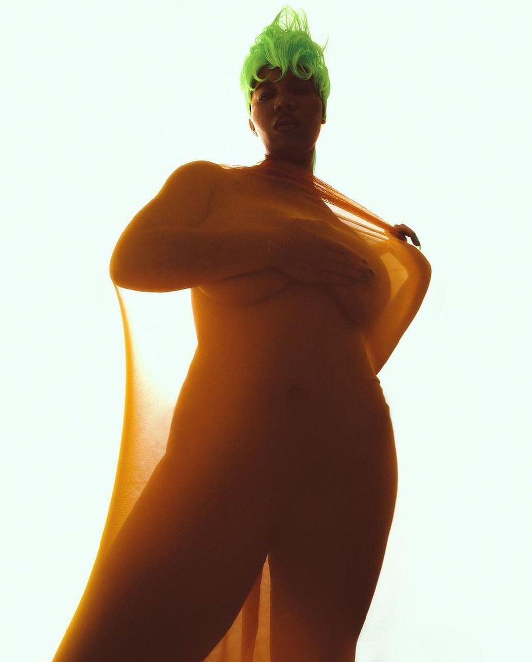 A woman with green hair poses in an orange bodysuit. The model stands in front of a bright background in a studio setting.