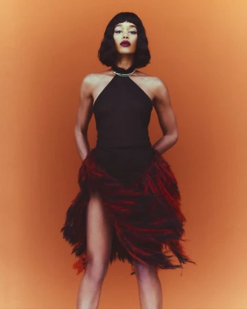 Elegant woman poses in a black halter dress with a feathered skirt against an orange backdrop in a fashion or beauty editorial shot.