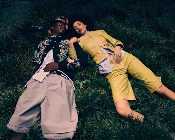 Two models lying on dark green grass, a black man in embroidered jacket and a woman in olive yellow coordinates.