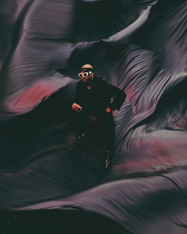 Adult male wearing sunglasses and a hat poses against a dark draped fabric background, creating a dramatic and artistic visual.